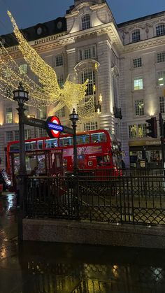 a red double decker bus parked in front of a building with christmas lights on it