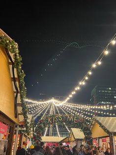 many people are walking through an outdoor market with lights on the buildings and trees in the background