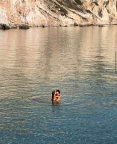 two people are swimming in the water near some hills and cliffs, one is holding his arms up