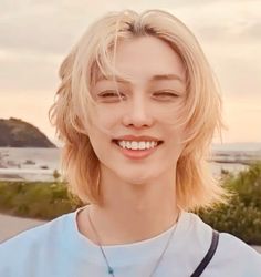 a woman with blonde hair smiling and wearing a white shirt in front of the ocean