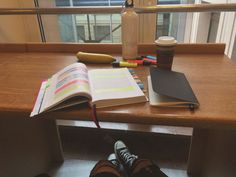 a person's feet resting on a desk with a book, coffee cup and pen