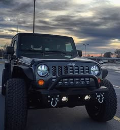 a jeep is parked in a parking lot with cloudy skies behind it and lights on