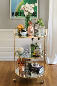 a gold bar cart filled with drinks on top of a hard wood floor next to a painting