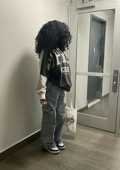 a woman with curly hair standing in front of a door holding a bag and looking at the wall