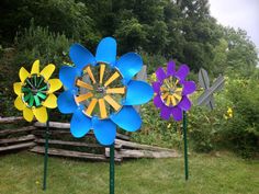 three colorful metal flowers sitting on top of green grass next to a wooden fence and trees