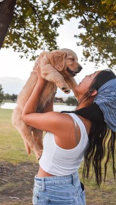 a woman holding a dog up to her face in the air while standing next to a tree