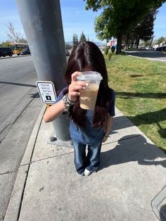 a woman holding up a plastic cup on the side of a street next to a pole