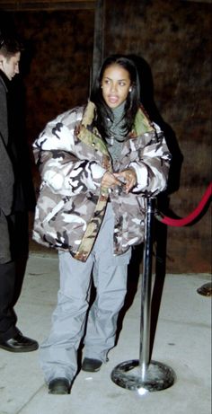 a woman standing in front of a pole wearing a camouflage jacket and scarf with her hands on her hips