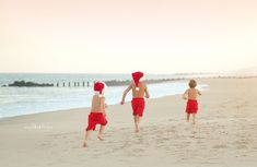 three children running on the beach in red trunks and santa's hats, with one child holding his hand