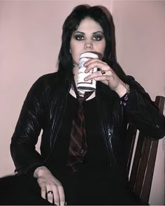 a woman sitting in a chair drinking from a coffee cup while wearing a leather jacket and tie