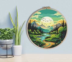 a cross - stitch landscape is displayed on a wall next to a potted plant