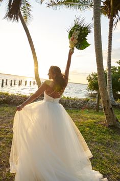 a woman in a white wedding dress holding flowers up to the sky with palm trees behind her