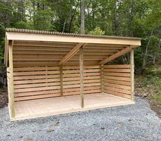 a wooden carport in the middle of a gravel area next to trees and bushes