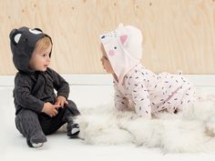 two toddlers dressed in animal onesuits sitting on a white rug and looking at each other