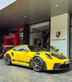 a yellow sports car parked in front of a porsche dealership with two motorcycles behind it