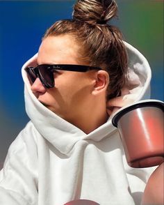 a woman wearing sunglasses and a hoodie holds a coffee cup in front of her face