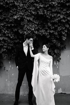 a man and woman standing next to each other in front of a plant covered wall