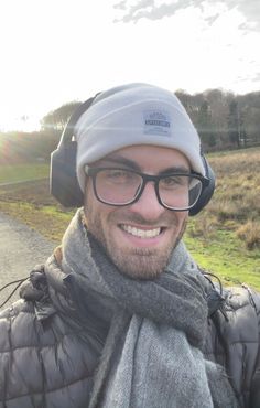 a man wearing glasses and a beanie smiles at the camera while standing on a road