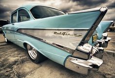 an old blue car parked in a parking lot under a cloudy sky with clouds above it