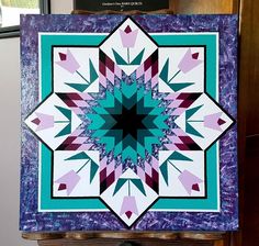 a purple and blue quilt with an intricate design on the front, sitting on top of a wooden table