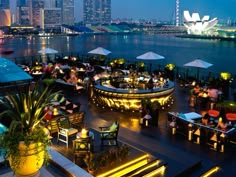 an outdoor dining area with tables and umbrellas overlooking the water at night, lit up by bright lights