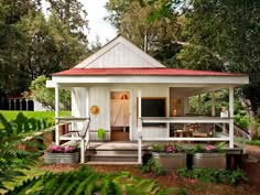 a small white house with a red roof in the middle of some trees and bushes