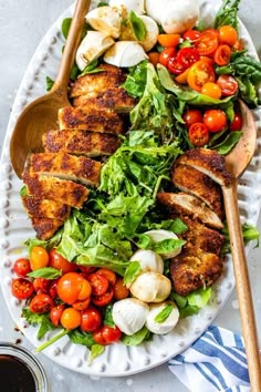 a white plate topped with chicken, tomatoes and lettuce next to a wooden spoon