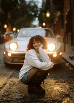 a woman squatting on the sidewalk in front of a car with her arms crossed