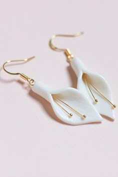 two white leaf shaped earrings with gold earwires on a pink surface, one is dangling