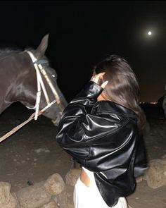 a woman wearing a black leather jacket is standing next to a brown horse at night