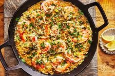 a skillet filled with shrimp and rice on top of a wooden table next to lemon wedges