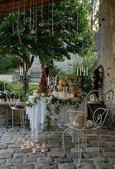 an outdoor dining table with candles and flowers on it, surrounded by greenery in front of a stone wall