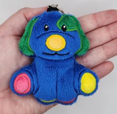 a hand holding a blue stuffed animal with yellow and green accents on it's face