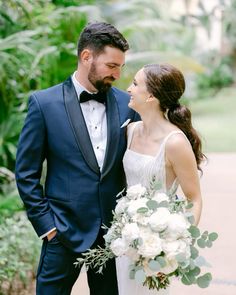 a bride and groom smile at each other as they stand in front of some greenery