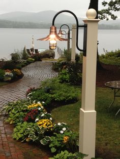 a light that is on the side of a pole near some flowers and plants in front of a body of water