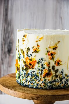 a cake with white frosting and colorful flowers on it sitting on a wooden stand