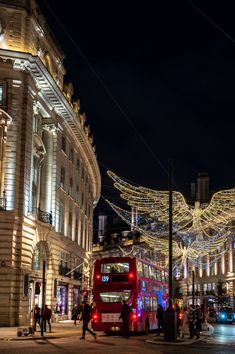 a red double decker bus driving down a street next to tall buildings covered in christmas lights