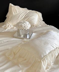 a white bed with ruffled pillows and a flower vase on it's side