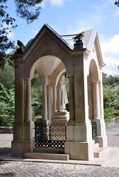 an outdoor gazebo with statues on the top and side walls, in front of trees