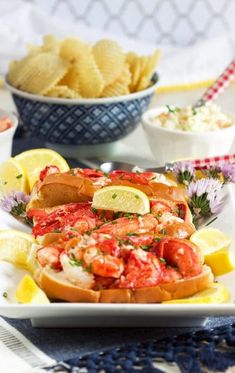 lobster sandwiches with lemon wedges and chips on a plate next to a bowl of coleslaw