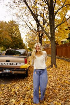 a woman standing in leaves next to a truck and trees with yellow leaves on the ground