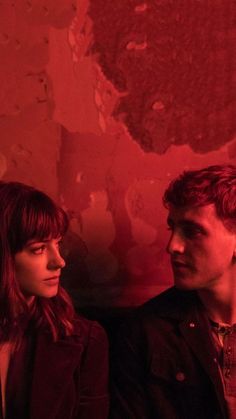 a man and woman standing next to each other in front of a red wall with peeling paint on it