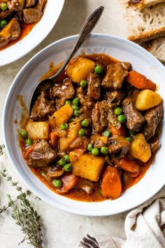 two white bowls filled with beef stew and potatoes on top of a table next to slices of bread