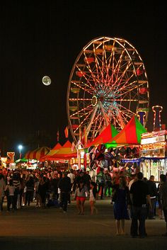 an amusement park at night with people walking around and ferris wheel in the background,