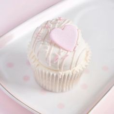 a white cupcake with pink frosting and a heart on top sitting on a plate