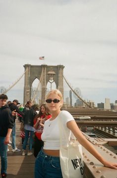 a woman standing on top of a bridge holding onto a white bag with the brooklyn bridge in the background