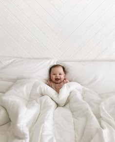 a baby is smiling while laying in a white comforter on a bed with pillows