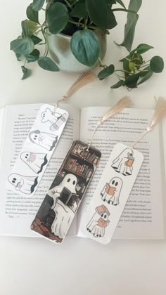 two bookmarks with ghost characters on them sitting next to a potted plant and an open book