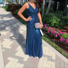 a woman in a long blue dress standing on a brick walkway next to flowers and palm trees