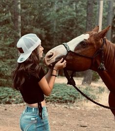 a woman in jeans and a white hat petting the head of a brown horse